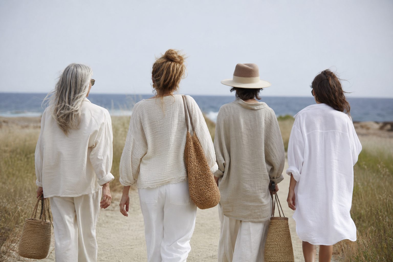 Small group of women in linen layers walking a bright coastal path above blue water